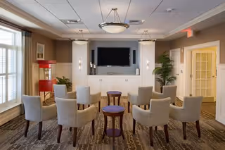 A senior living facility common area with eight beige upholstered chairs arranged in two rows facing a large flat-screen TV mounted on a wall. The room has neutral-colored walls, carpeted flooring, two small round wooden tables, a popcorn machine in the corner, and large windows with curtains allowing natural light to enter.