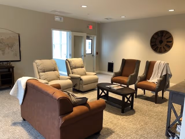 A cozy living room area with a brown leather sofa, two beige recliners, and two brown armchairs arranged around a dark wooden coffee table. The room has beige walls, a carpeted floor, a large round wall clock, and a door leading outside with a window beside it.