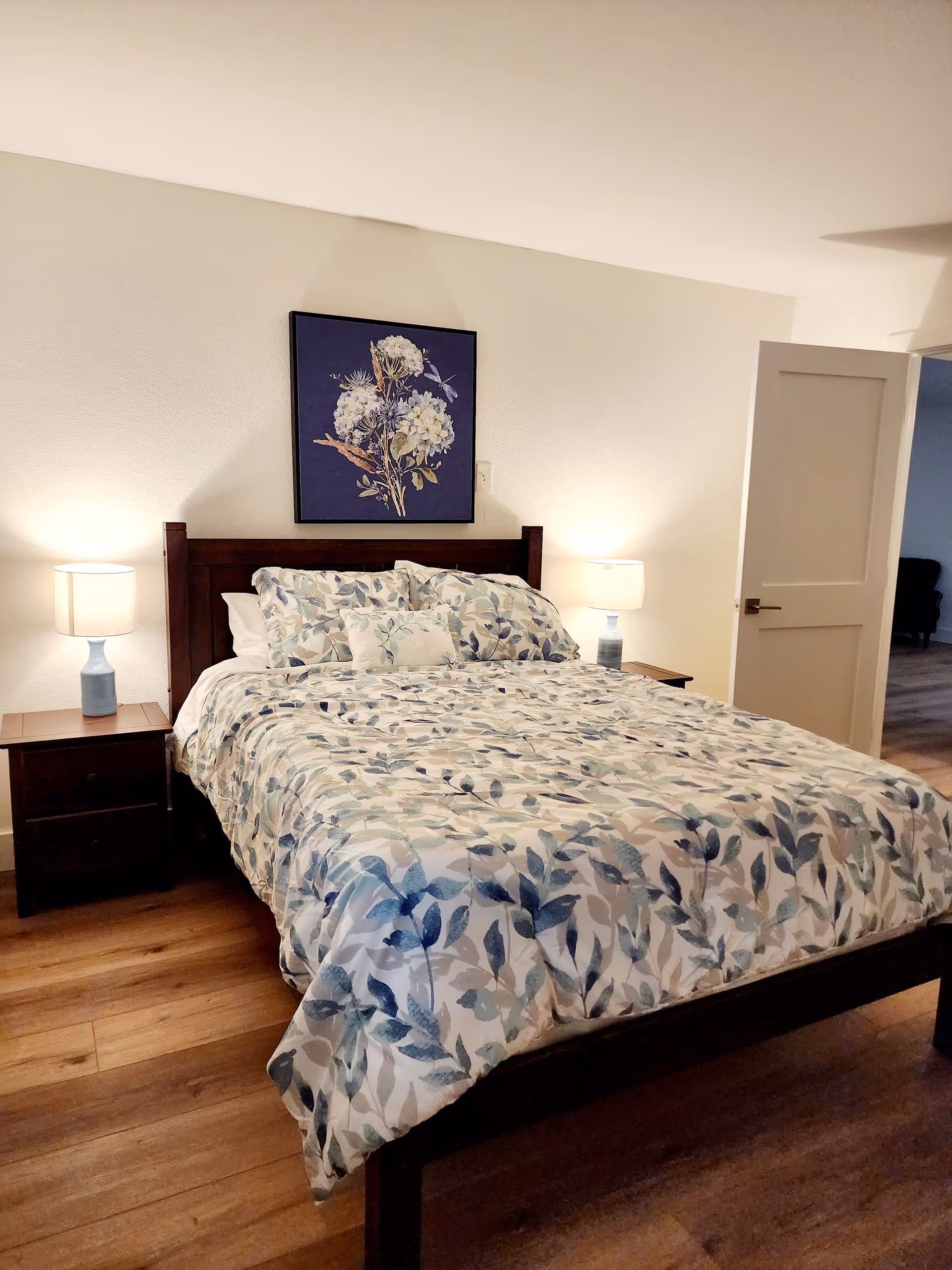 A bedroom with a wooden bed frame and a bed made with a floral patterned comforter and pillows in shades of blue and beige. There are two wooden nightstands on either side of the bed, each with a blue lamp with a white lampshade. Above the bed hangs a framed artwork featuring white flowers on a dark blue background. The room has wooden flooring and white walls, with an open door leading to another room.
