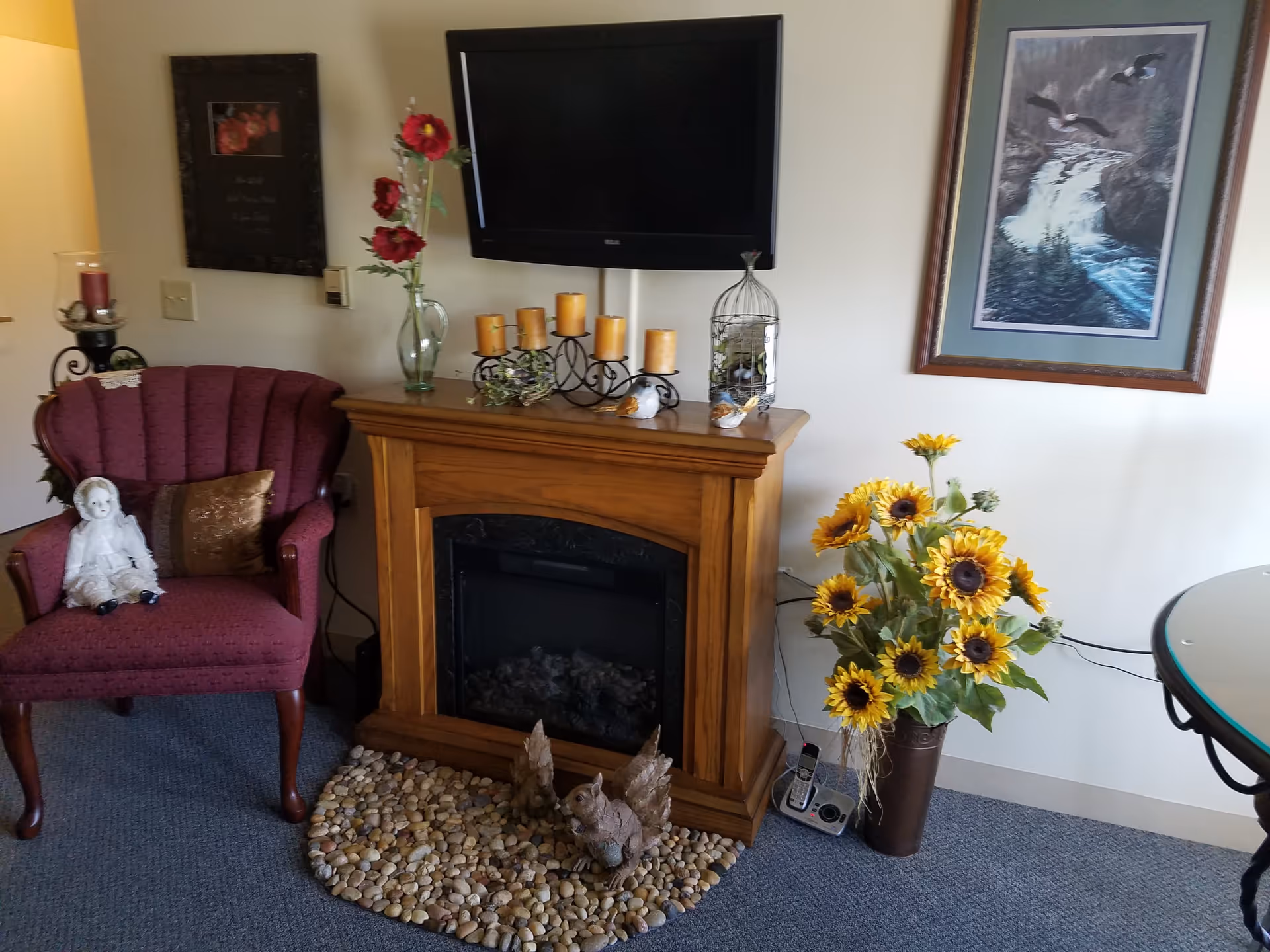 A cozy living room corner featuring a burgundy upholstered armchair with a decorative doll sitting on it, a wooden electric fireplace with candles and decorative items on top, a flat-screen TV mounted above the fireplace, a vase with red flowers, a large bouquet of sunflowers in a brown vase, a framed picture of ducks flying over a waterfall on the wall, and a round glass table partially visible on the right.