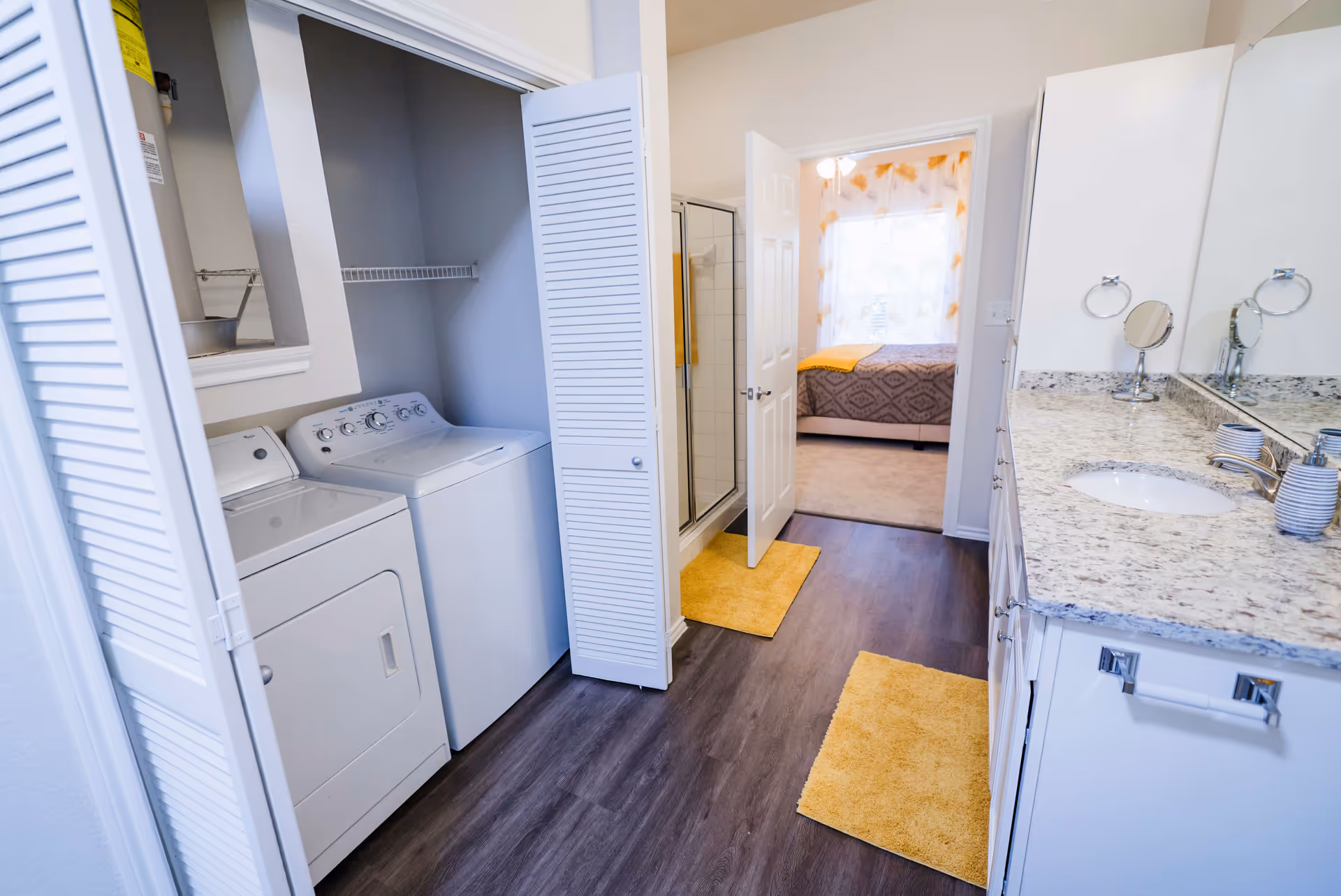 Interior view of a laundry area with a washer and dryer behind white louvered doors, adjacent to a bathroom with a granite countertop, sink, and mirror. The bathroom has yellow rugs on the floor and a glass shower door. Beyond the bathroom, a bedroom with a bed covered in a patterned bedspread and yellow accents is visible through an open door.