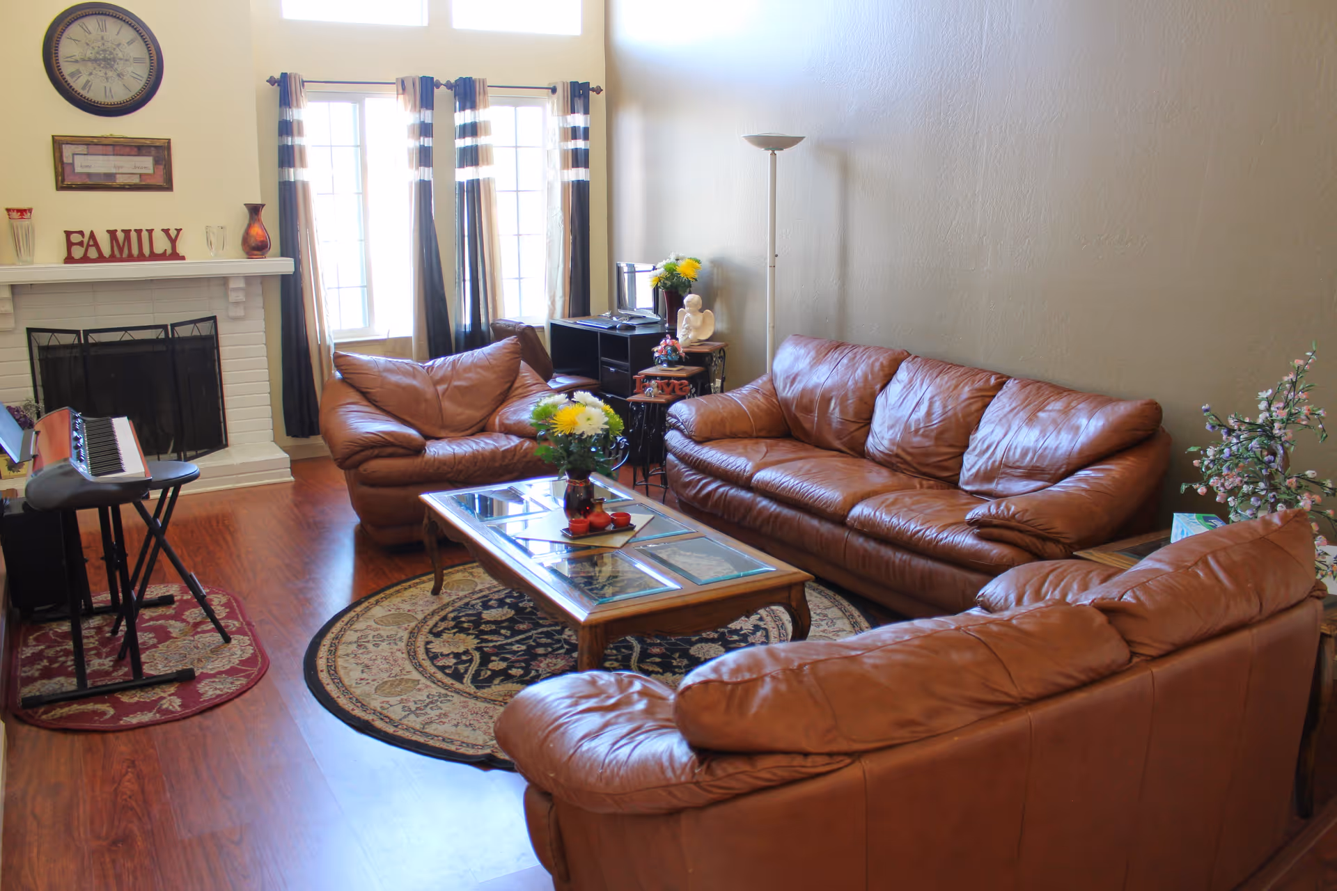 A cozy living room with three brown leather sofas arranged around a wooden coffee table with a glass top. The room features a fireplace with the word FAMILY displayed on the mantel, a round rug under the coffee table, a keyboard on a stand, a floor lamp, and a window with striped curtains letting in natural light.