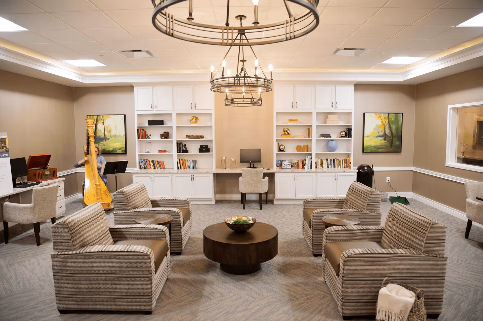 A bright senior living lounge with four striped armchairs around a round coffee table, built-in bookshelves, and a harp in the corner.