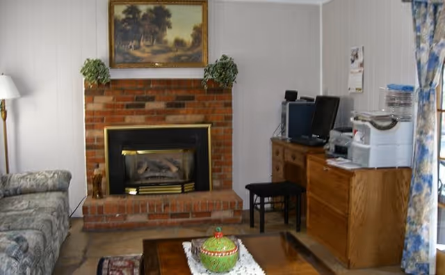 A cozy living room with a brick fireplace in the center, a framed landscape painting above it, a patterned sofa on the left, a wooden coffee table with a decorative green jar in the middle, and a wooden dresser with office equipment including a computer and printer on the right. Floral curtains cover a window on the far right.
