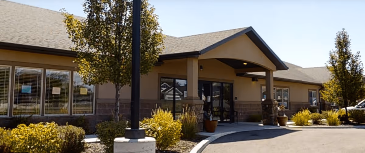 Exterior view of a single-story assisted living facility building with a covered entrance, large windows, and landscaped bushes and trees in front under a clear sky.