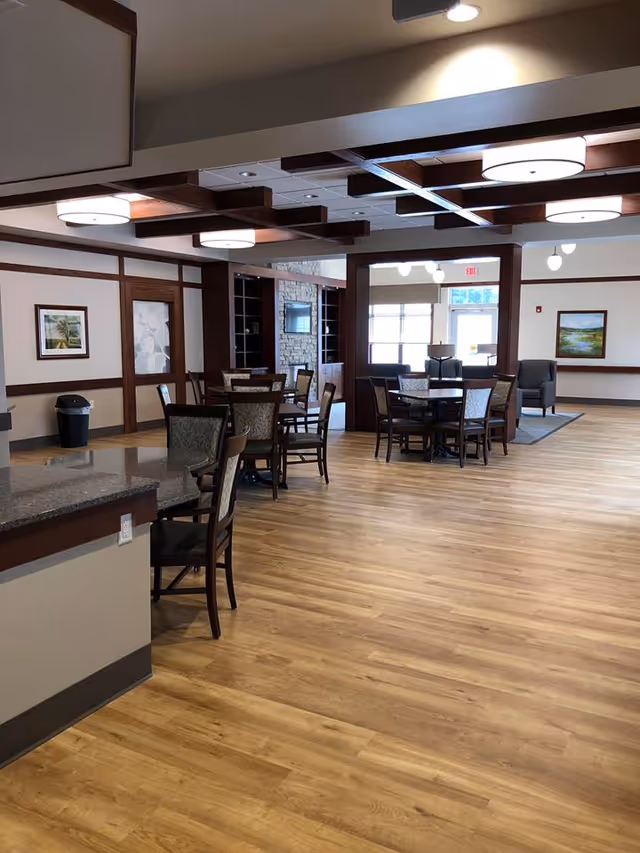 Spacious interior room with wooden flooring and ceiling beams, featuring several round tables with chairs arranged around them. The room has large windows allowing natural light, framed artwork on the walls, and a stone fireplace with a mounted TV in the background. There is also a countertop with chairs in the foreground.