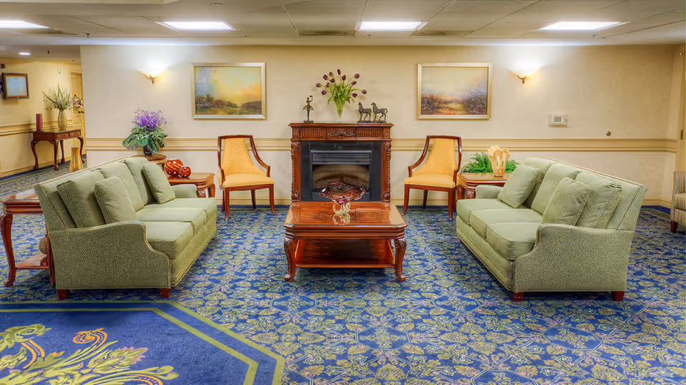 A cozy living room area with two green upholstered sofas facing each other, a wooden coffee table in the center with a glass vase, two yellow cushioned chairs near a decorative fireplace, floral arrangements, and framed landscape paintings on the wall.