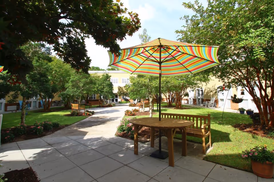 Sunlit courtyard with a striped patio umbrella over a wooden table and benches, paved walkways, trees, flower beds and the surrounding building in the background.
