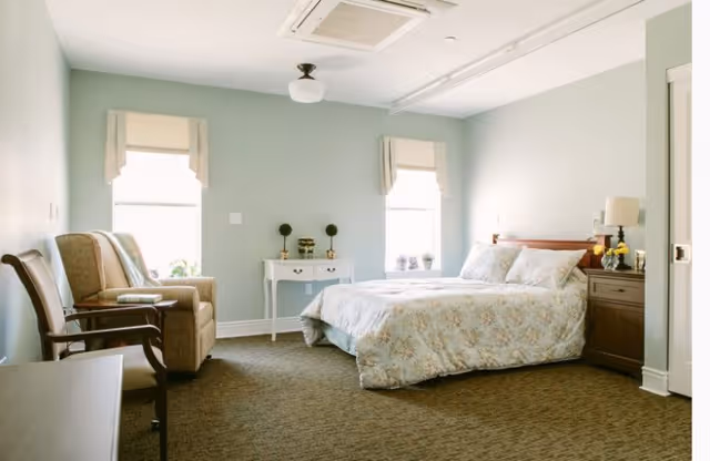 A bright and cozy bedroom with light green walls, two windows with beige valances, a bed with floral bedding, a wooden nightstand with a lamp and flowers, a small white table with decorative items, and two chairs near the wall.