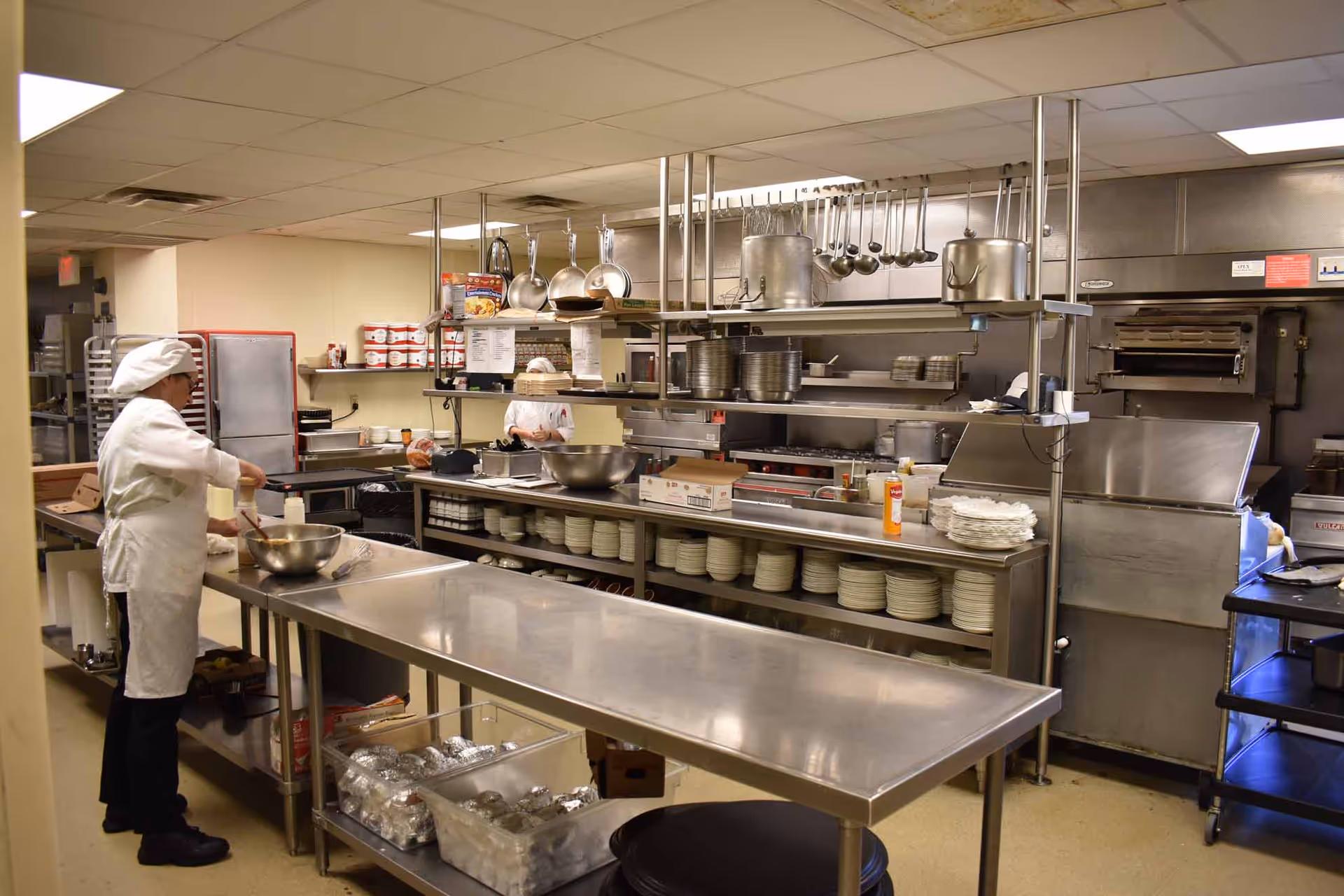 A commercial kitchen with stainless steel counters and shelves filled with plates, pots, and pans. Two chefs in white uniforms and hats are preparing food. Various kitchen equipment and supplies are visible, including an oven and containers of ingredients.