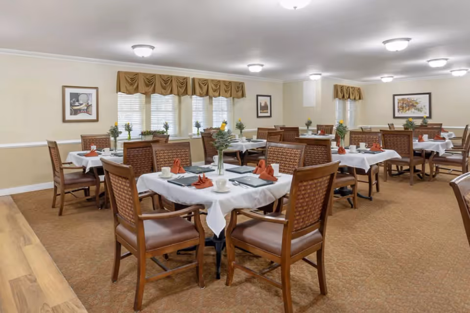 A dining room with multiple tables covered in white tablecloths, each set with cups, plates, napkins, and small flower vases. The room has beige walls, several windows with valances, framed artwork on the walls, and ceiling lights providing illumination.