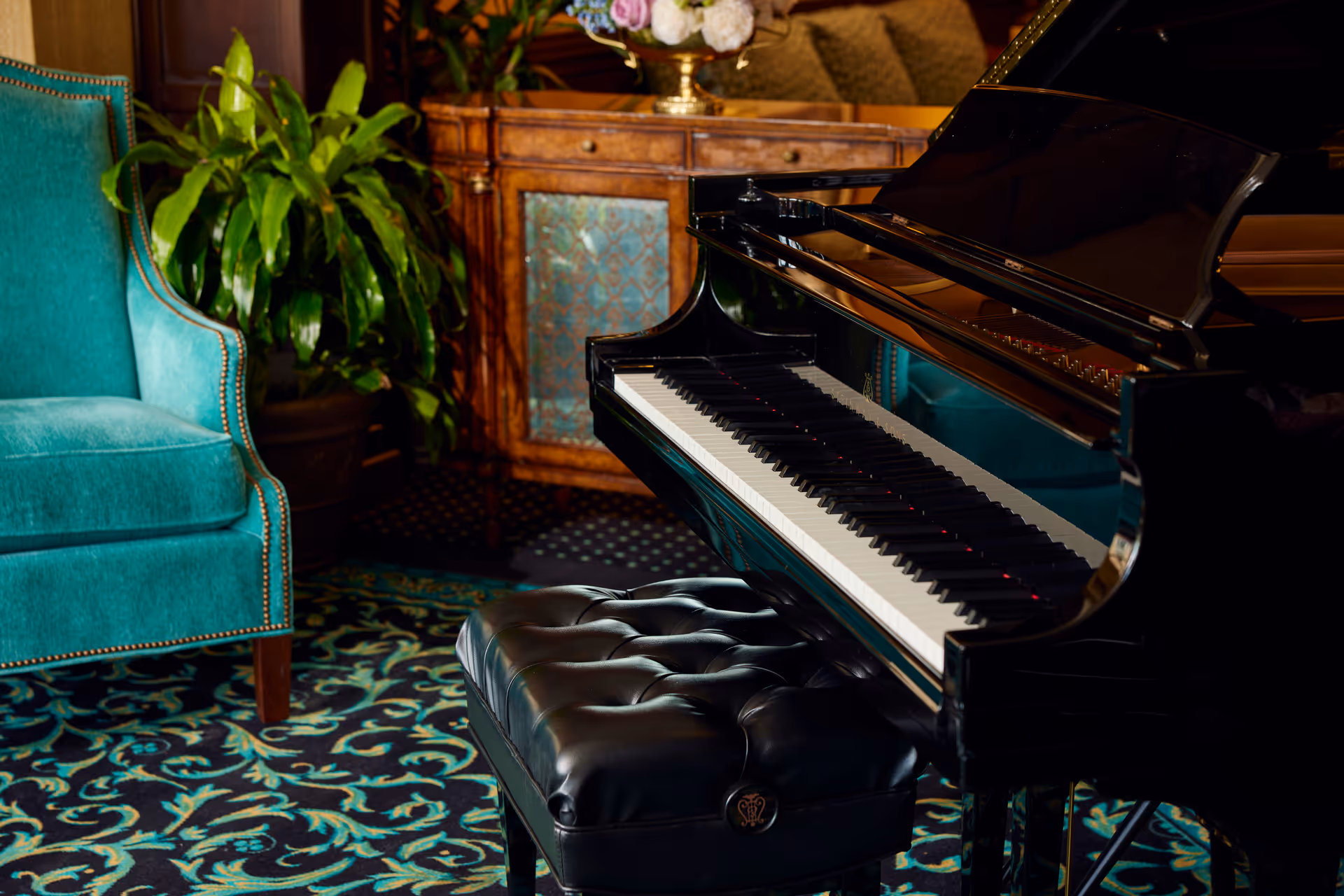 Black grand piano with a tufted bench next to a teal armchair, plant, and decorative cabinet in a patterned-carpet sitting area.