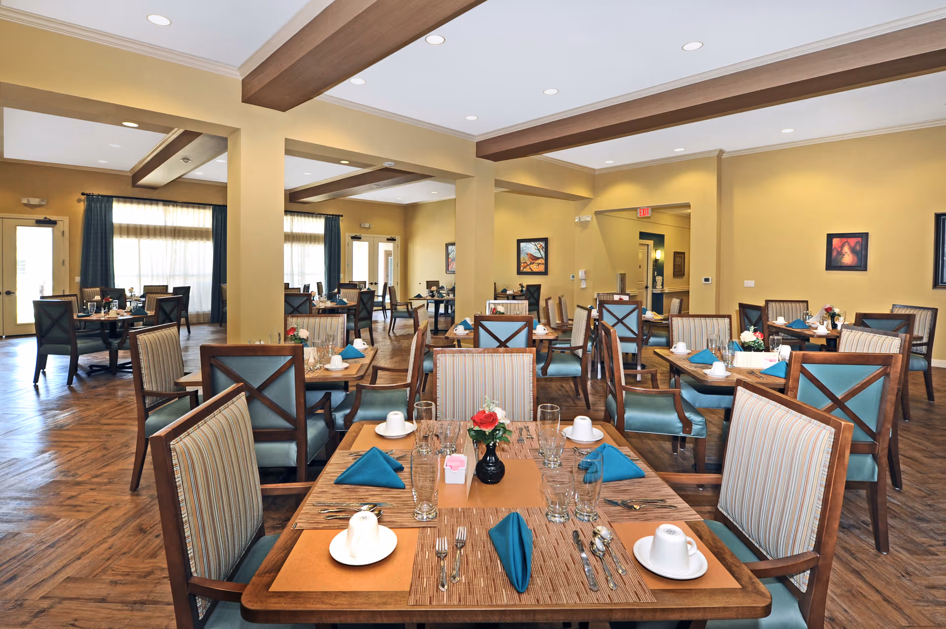 A spacious senior living dining room with multiple wooden tables and chairs arranged neatly. Each table is set with white cups, glasses, silverware, and teal cloth napkins. The room has warm yellow walls, wooden flooring, and large windows with curtains allowing natural light to enter. There are framed artworks on the walls and ceiling lights providing additional illumination.