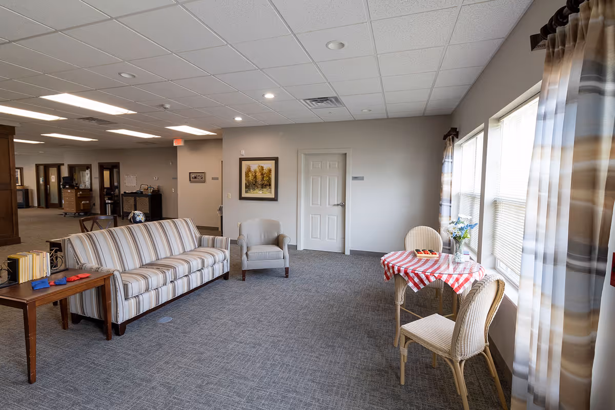 A bright and spacious common area in an assisted living facility featuring a striped sofa, a gray armchair, a small table with a red and white checkered tablecloth, two chairs, and large windows with striped curtains letting in natural light. The room has gray carpet and neutral-colored walls with a framed painting and a closed door.