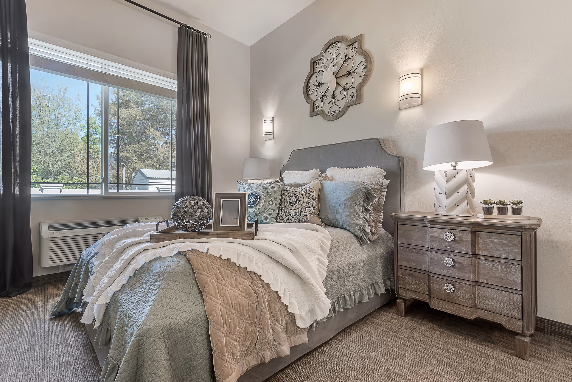 Bright, tastefully decorated bedroom with a made bed, patterned pillows, a wooden nightstand and lamp, wall sconces, and a large window.