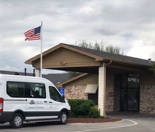 Front entrance of a care facility with a covered drive-through, an American flag on a pole, and a white passenger van parked nearby.