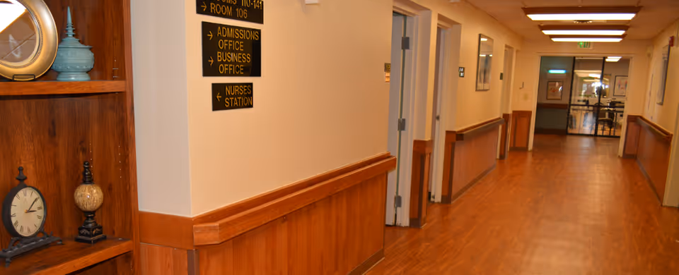 A well-lit hallway in a nursing and rehabilitation facility with wooden flooring and wood paneling on the lower half of the walls. There are several doors along the hallway, a wooden shelf with decorative items including a clock and vases on the left, and directional signs indicating rooms, admissions office, business office, and nurses station. The hallway leads to a glass door at the far end.