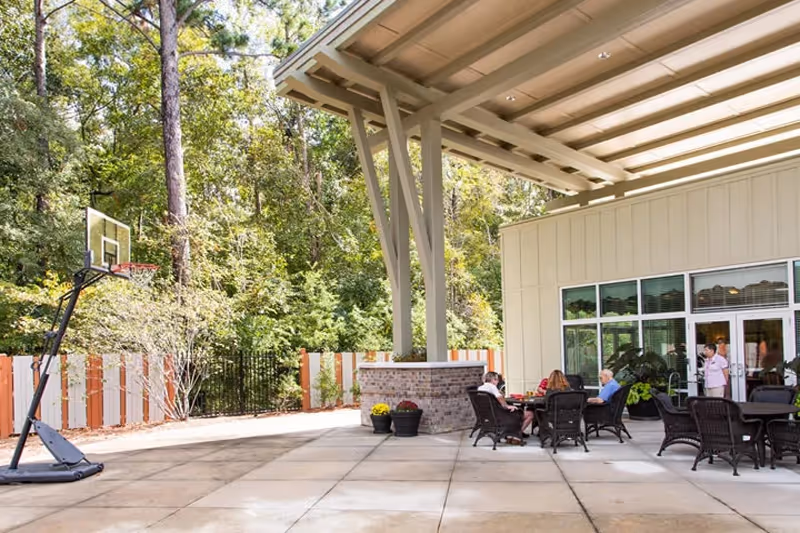 Outdoor covered patio area with several people sitting around tables and chairs. There is a portable basketball hoop on the left side and a wooded area with trees and a fence in the background.