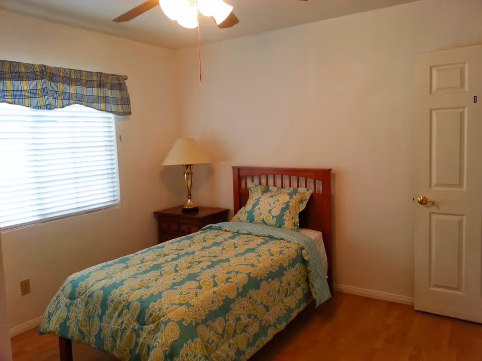 A small bedroom with a single bed covered in a blue and yellow floral patterned comforter and matching pillow. Next to the bed is a wooden nightstand with a brass lamp. A window with closed blinds and a blue and yellow plaid valance is on the left wall. The room has light-colored walls, a ceiling fan with a light, and a wooden floor.