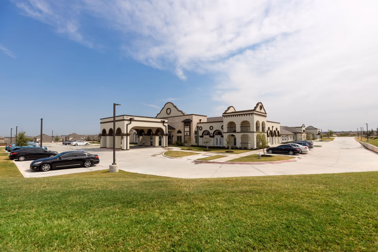 Front exterior of a Mediterranean-style senior living building with an arched portico, parking lot, and grassy foreground.