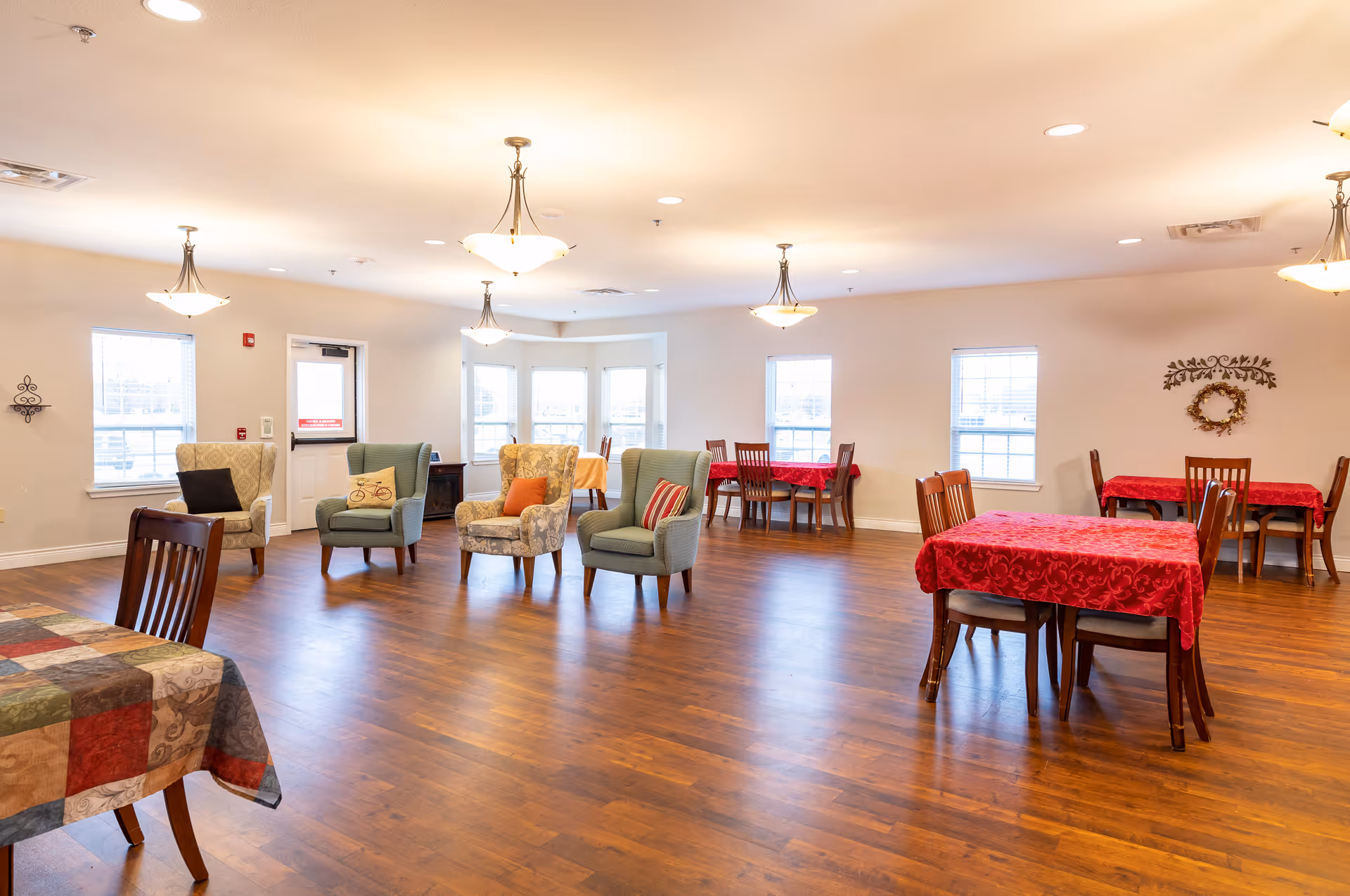 A spacious, well-lit common area with wooden flooring featuring several tables covered with red and patterned tablecloths, surrounded by wooden chairs. There are also four upholstered armchairs with decorative pillows arranged near the center of the room. The walls are light-colored with multiple windows allowing natural light to enter. Ceiling lights hang evenly spaced across the ceiling.