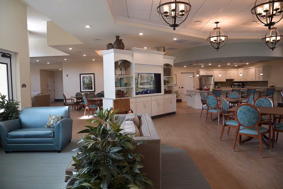 Open communal living and dining area with teal armchairs, round dining tables, a central white shelving divider, pendant lights, and a kitchen counter in the back.