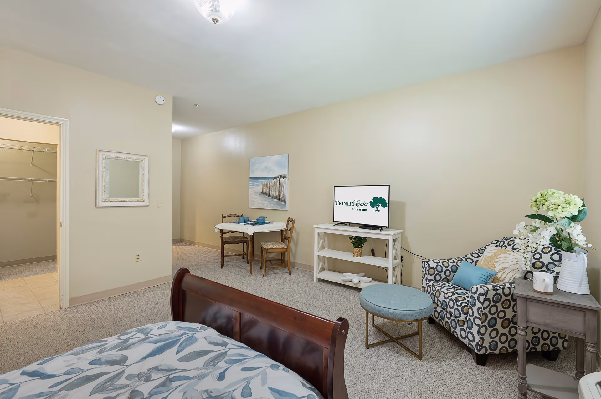 A senior living facility room with a bed in the foreground, a small dining table with two chairs, a patterned armchair with a blue pillow, a round ottoman, a side table with flowers, and a TV on a white stand displaying the Trinity Oaks of Pearland logo. There is an open doorway leading to a closet with wire shelving.