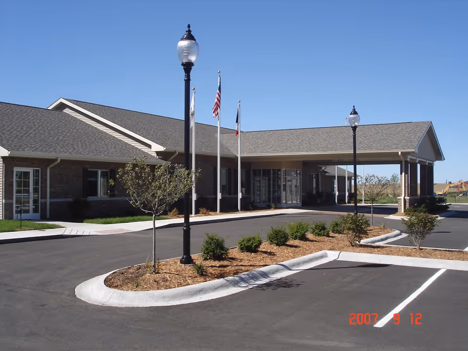 Front exterior of a senior living facility showing a covered entrance/porte-cochere, flagpoles, lampposts, and landscaped parking area.
