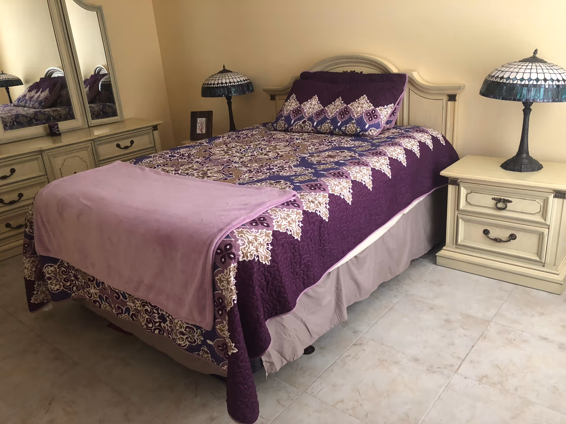 A neatly made bed with a purple and beige patterned quilt and matching pillow in a bedroom. The bed has a light purple blanket folded at the foot. On the right side of the bed is a cream-colored nightstand with two drawers and a decorative lamp with a stained glass shade. On the left side, there is a dresser with a large mirror reflecting part of the bed and lamp. The room has light-colored tiled flooring and beige walls.