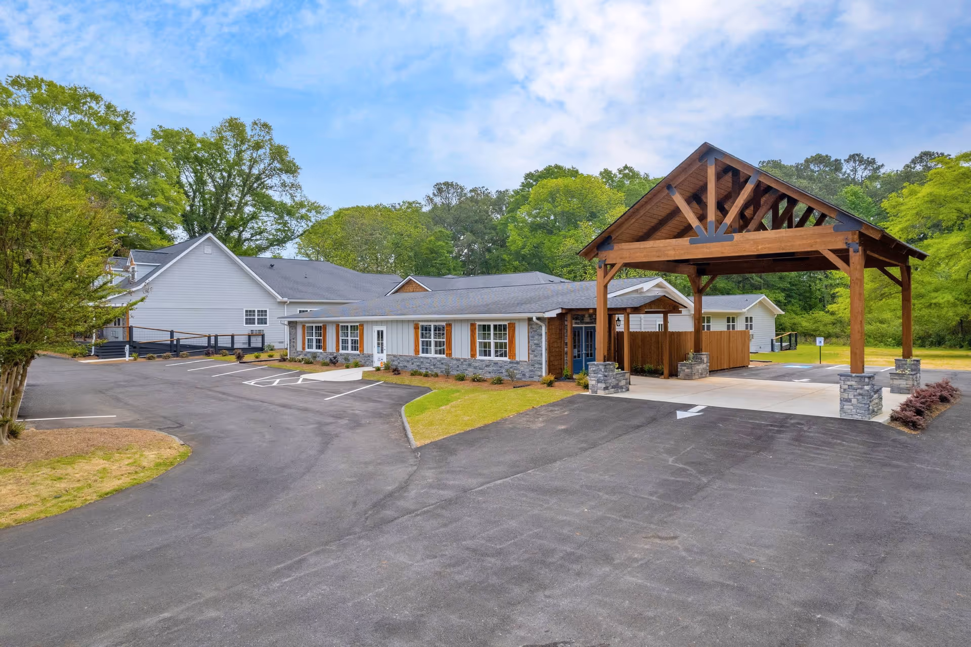Exterior view of The Joy Senior Living of Loganville showing a single-story building with a covered entrance supported by wooden beams and stone pillars. The building has white siding with stone accents and wooden shutters on the windows. There is a paved driveway and parking area with marked spaces, surrounded by green trees and grass under a partly cloudy blue sky.