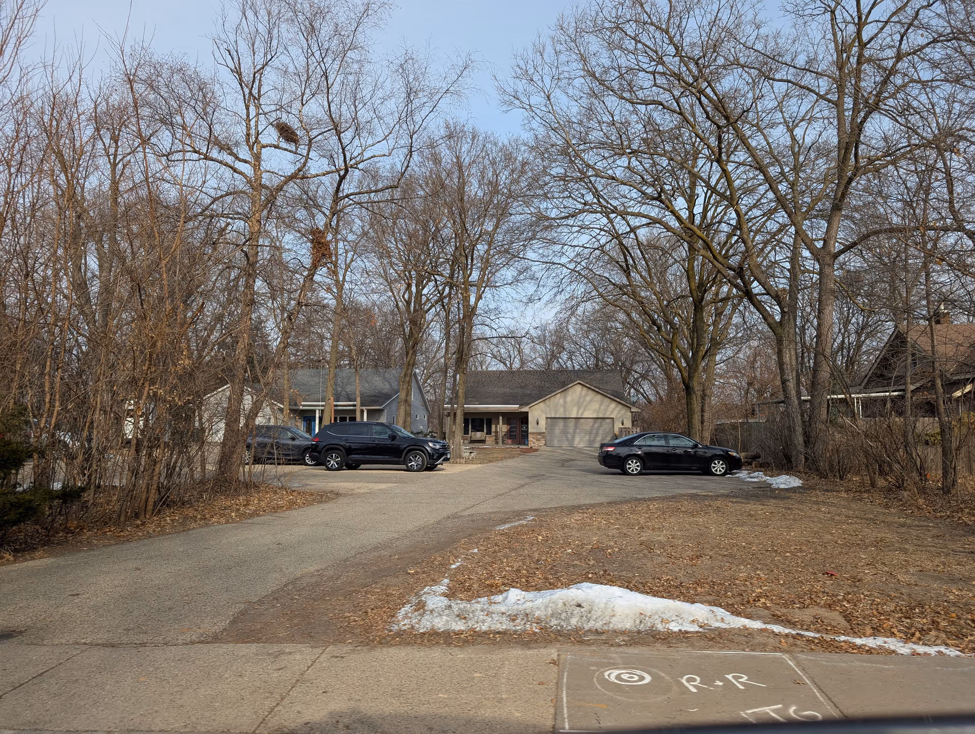 View of a residential care center building with a driveway and parked cars in front. Leafless trees surround the area, and patches of snow are visible on the ground. The sky is clear and blue.
