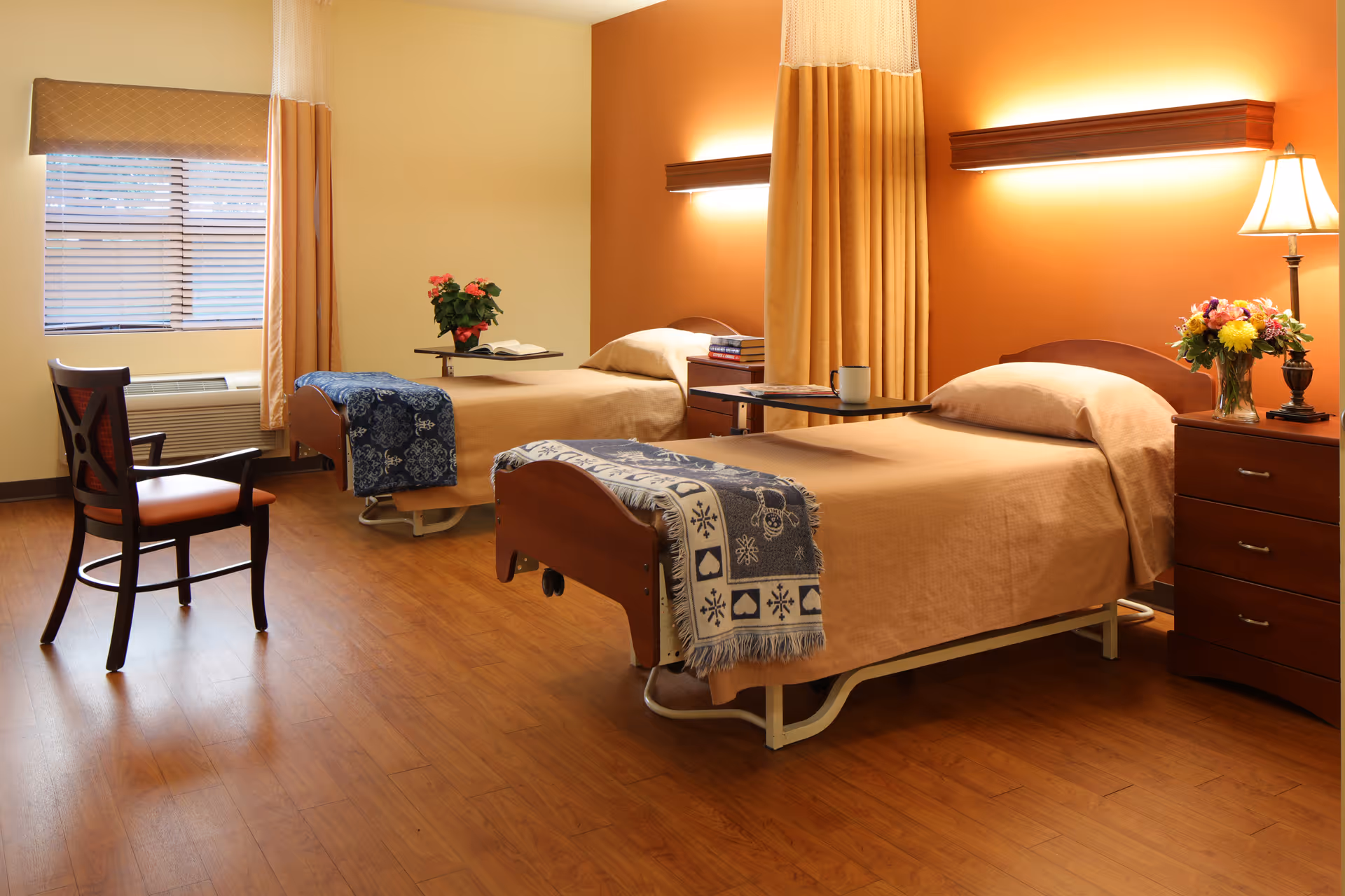 Two neatly made twin beds in a warm-toned nursing facility room with wooden flooring, bedside tables, a chair, and flowers.