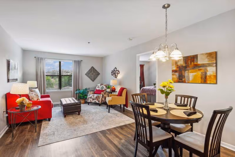 Open living and dining room with a red sofa and patterned chairs, a round dining table set under a chandelier, and a window letting in natural light.