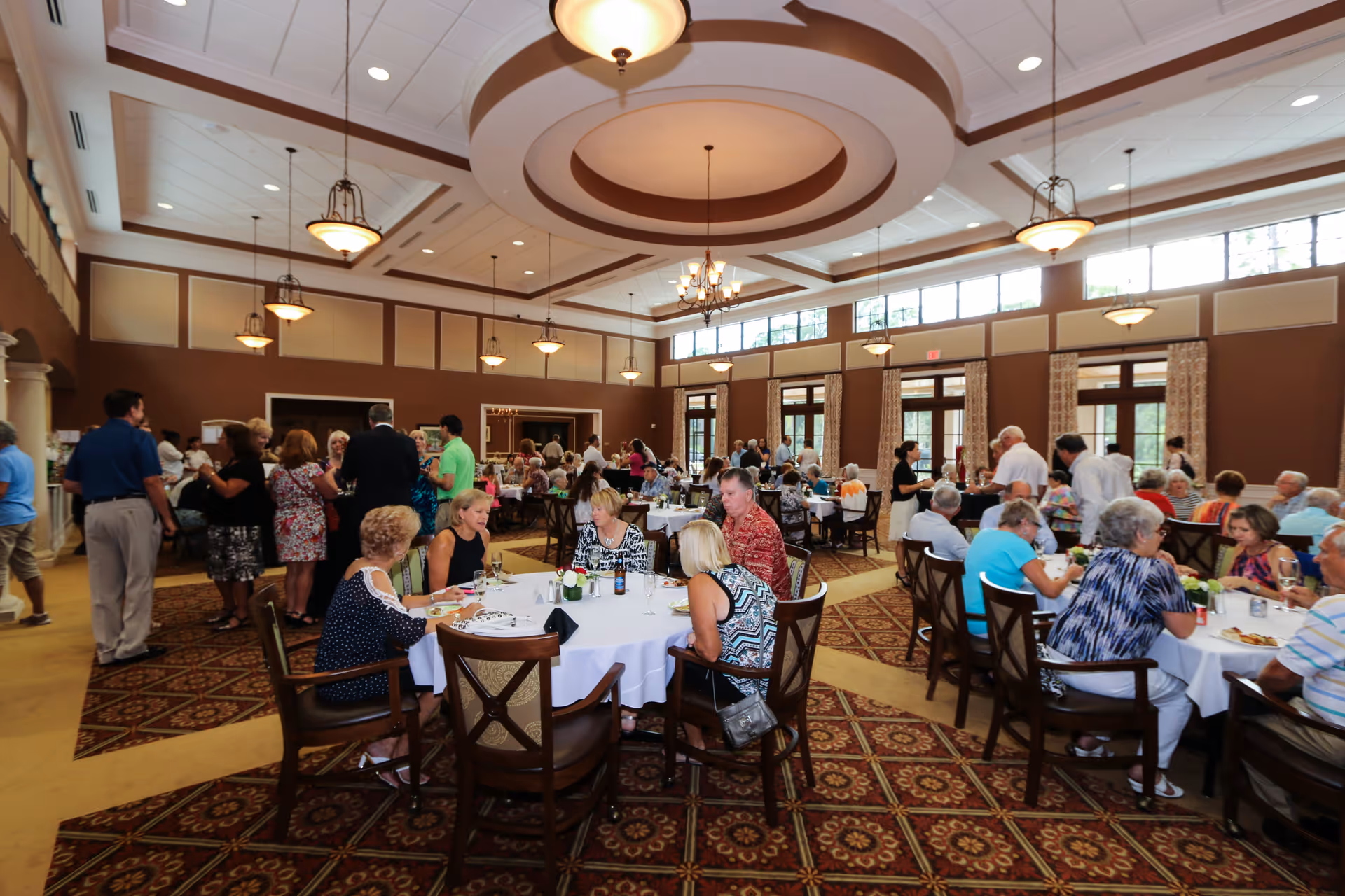 A large dining room filled with senior residents and staff socializing and dining at round tables covered with white tablecloths. The room features high ceilings with decorative light fixtures, large windows with curtains, and a patterned carpet. People are engaged in conversation and enjoying a communal meal in a bright, spacious setting.