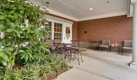 Covered outdoor seating area at a senior living entrance with metal chairs and table, cushioned benches, brick wall, and plants.