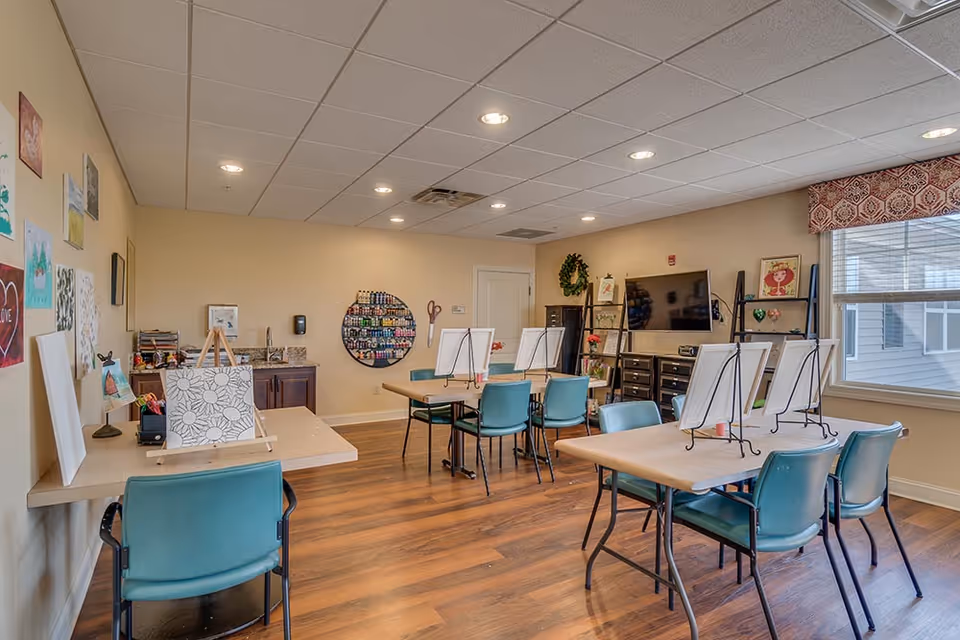 Bright activity/craft room with tables, easels, teal chairs, art supplies, and a TV on the wall.