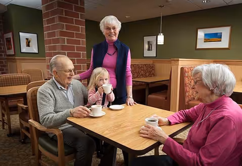 Three elderly adults and a young girl sitting and standing around a wooden table in a dining area, drinking from white cups. The room has green walls, framed pictures, and booth seating with patterned cushions.