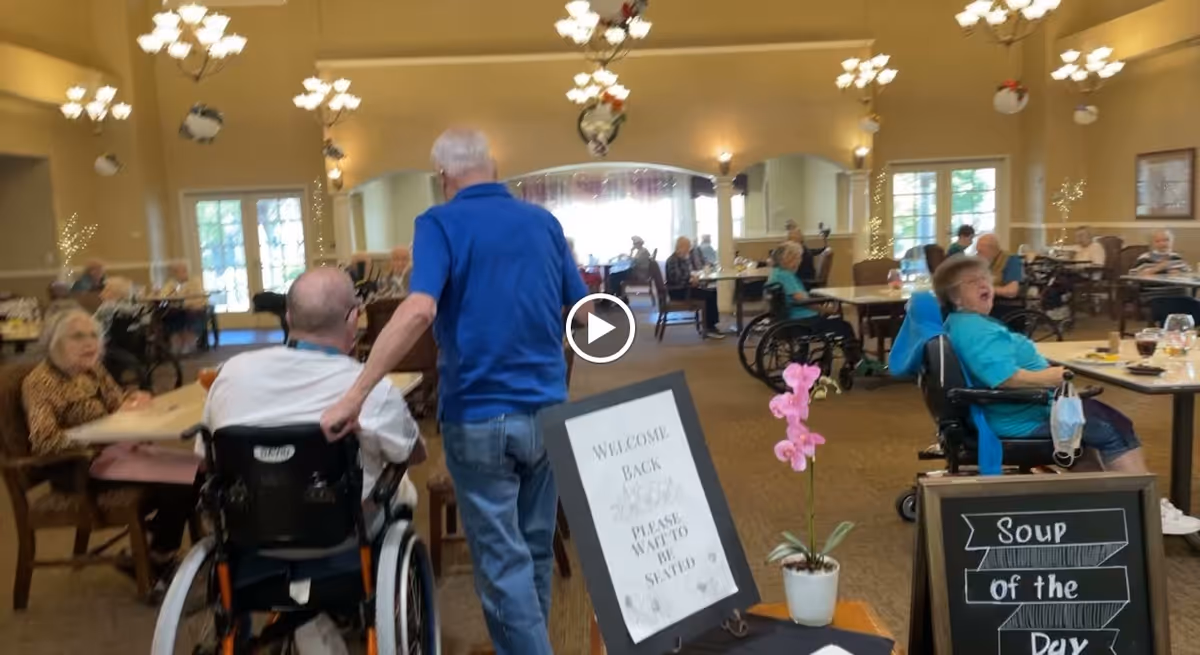 A dining room in a senior living facility with elderly residents seated at tables, some in wheelchairs. A man in a blue shirt is pushing another man in a wheelchair. The room is warmly lit with chandeliers and decorated with hanging ornaments. There are signs on stands, one welcoming guests and another displaying the soup of the day. Large windows let in natural light.