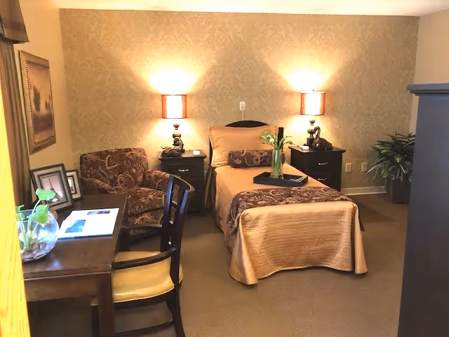 A furnished bedroom with a single bed flanked by two bedside tables and lamps, an armchair, and a desk.