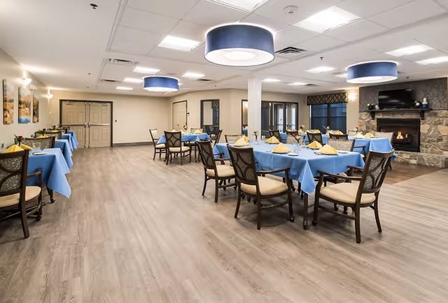 A spacious dining room with multiple tables covered in blue tablecloths and set with yellow napkins and glassware. The room features wooden chairs with cushioned seats, a stone fireplace with a TV mounted above it, and modern ceiling lights with blue shades. The floor is light wood, and there are paintings on the wall and large windows with dark blinds.