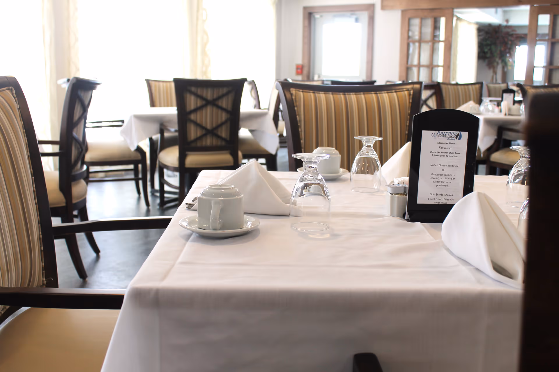 A dining area in a senior living facility with tables covered in white tablecloths, set with upside-down wine glasses, white napkins folded in a triangular shape, cups on saucers, and a menu stand. The chairs have striped upholstery and dark wooden frames. Large windows in the background let in natural light.