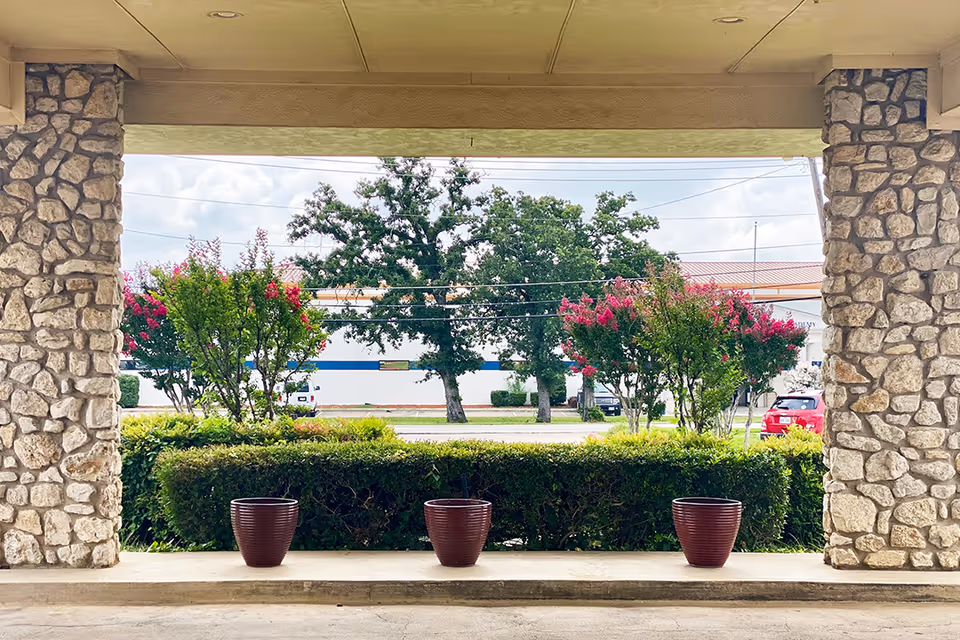 View from a covered stone patio area looking out onto a landscaped garden with green bushes and flowering trees. Beyond the garden, there is a street with parked vehicles and a building in the background under a partly cloudy sky.
