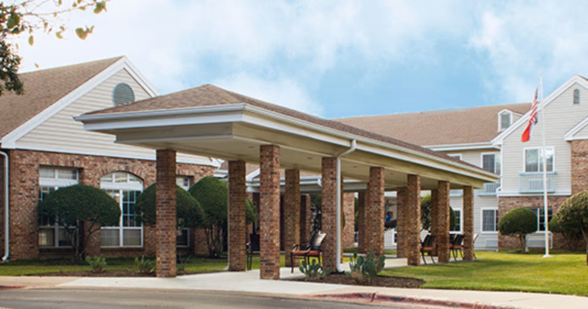 Exterior view of Juniper Village at Spicewood Summit showing a covered entrance supported by brick columns, manicured bushes, and a flagpole with an American flag in front of a two-story building with brick and siding.