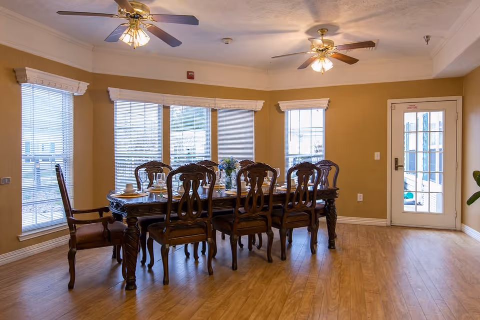 A dining room with a large wooden dining table and eight matching chairs. The table is set with plates, cups, and a floral centerpiece. The room has wooden flooring, beige walls, three ceiling fans with lights, and several windows with blinds. A glass door is visible on the right side of the room.