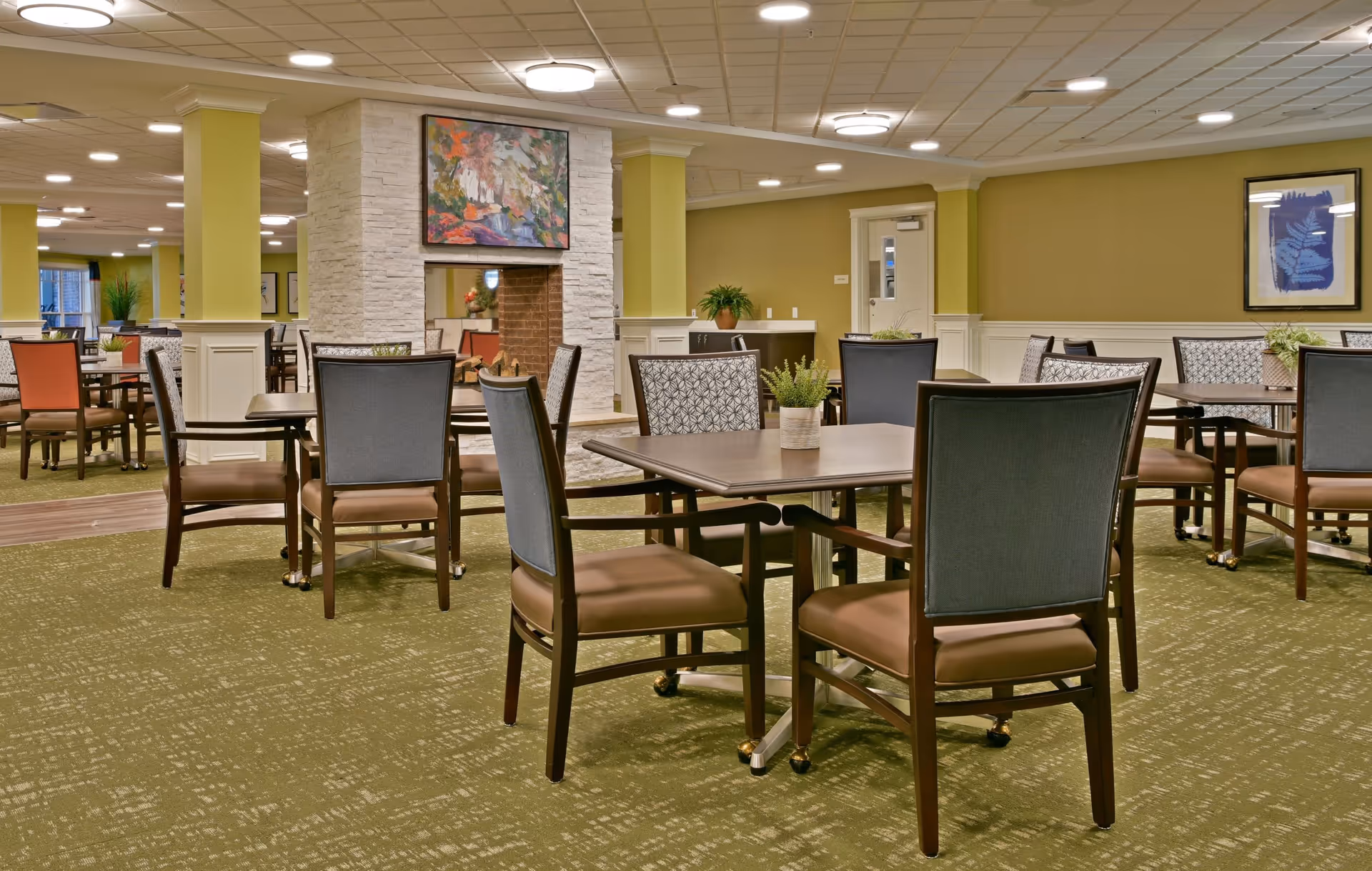 A spacious dining room in a senior living facility with multiple tables and chairs arranged neatly. The room features green carpeting, light green walls with white wainscoting, and a central fireplace with a colorful painting above it. There are potted plants on some tables and framed artwork on the walls.