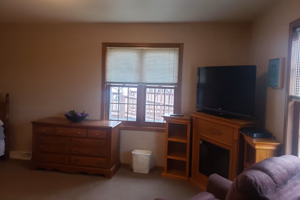 A cozy senior living room with a wooden dresser topped with a flower arrangement, a window with blinds partially closed, a flat-screen TV on a wooden fireplace mantel, and a comfortable upholstered chair in the foreground.
