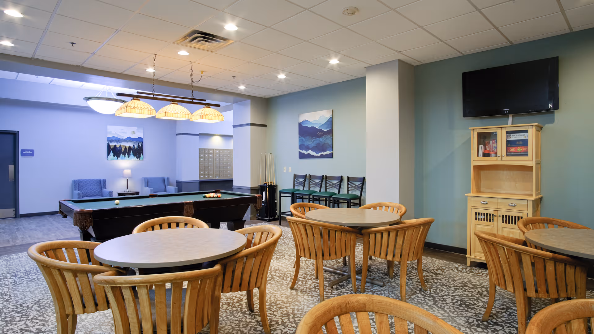 A senior living community common room with round tables and chairs, a pool table, TV, and a seating area.