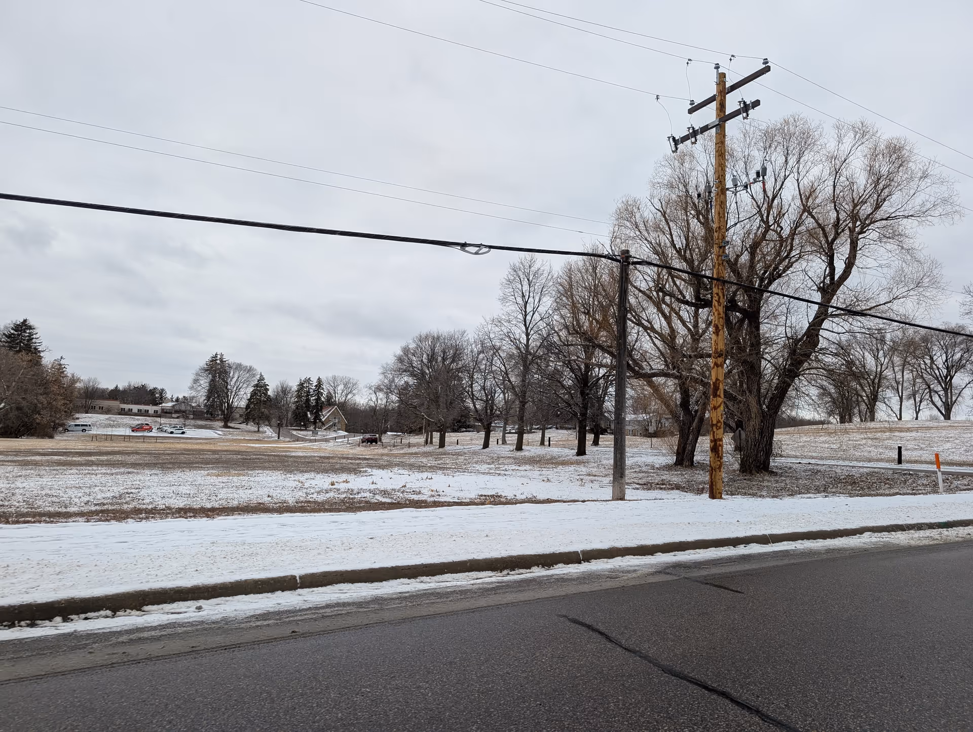 A snowy landscape with a road in the foreground, utility poles and power lines, leafless trees, and a few houses and vehicles in the distance under a cloudy sky.