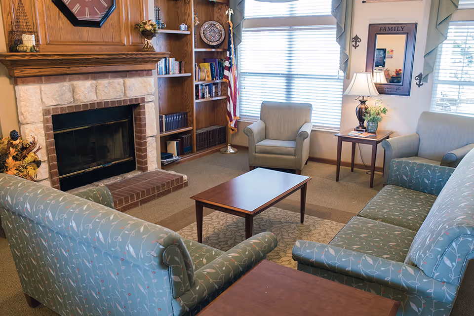 A cozy living room with a stone fireplace, wooden mantel, and built-in bookshelves. The room features two patterned sofas, two armchairs, a wooden coffee table, a side table with a lamp and flowers, and large windows with blinds letting in natural light. An American flag stands in the corner, and a mirror with the word 'FAMILY' hangs on the wall.