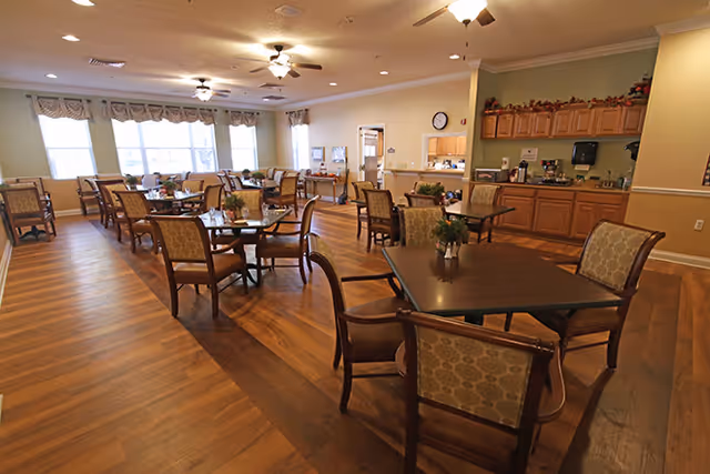 A spacious dining room with multiple wooden tables and cushioned chairs arranged neatly. The room has large windows with valances allowing natural light to fill the space. There is a counter area with cabinets and coffee machines along one wall. The floor is wood with a darker wood inlay design, and ceiling fans with lights are mounted on the ceiling.
